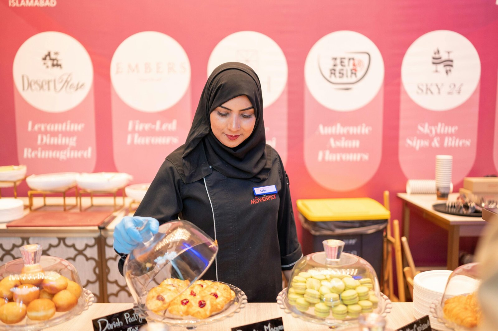 Female chef in hijab serves pastries at a stylish bakery in Islamabad, showcasing delectable treats.