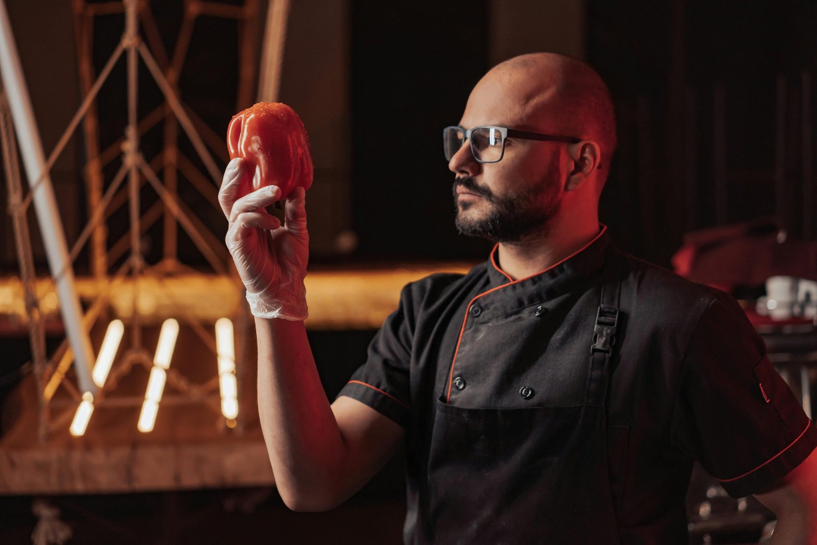 Chef in a dark kitchen holding a red bell pepper, showcasing culinary expertise.